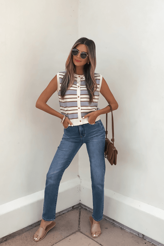 Woman in a Multi Blue Striped Sweater Vest, sunglasses, blue jeans, sandals, and a brown shoulder bag stands by a white wall.
