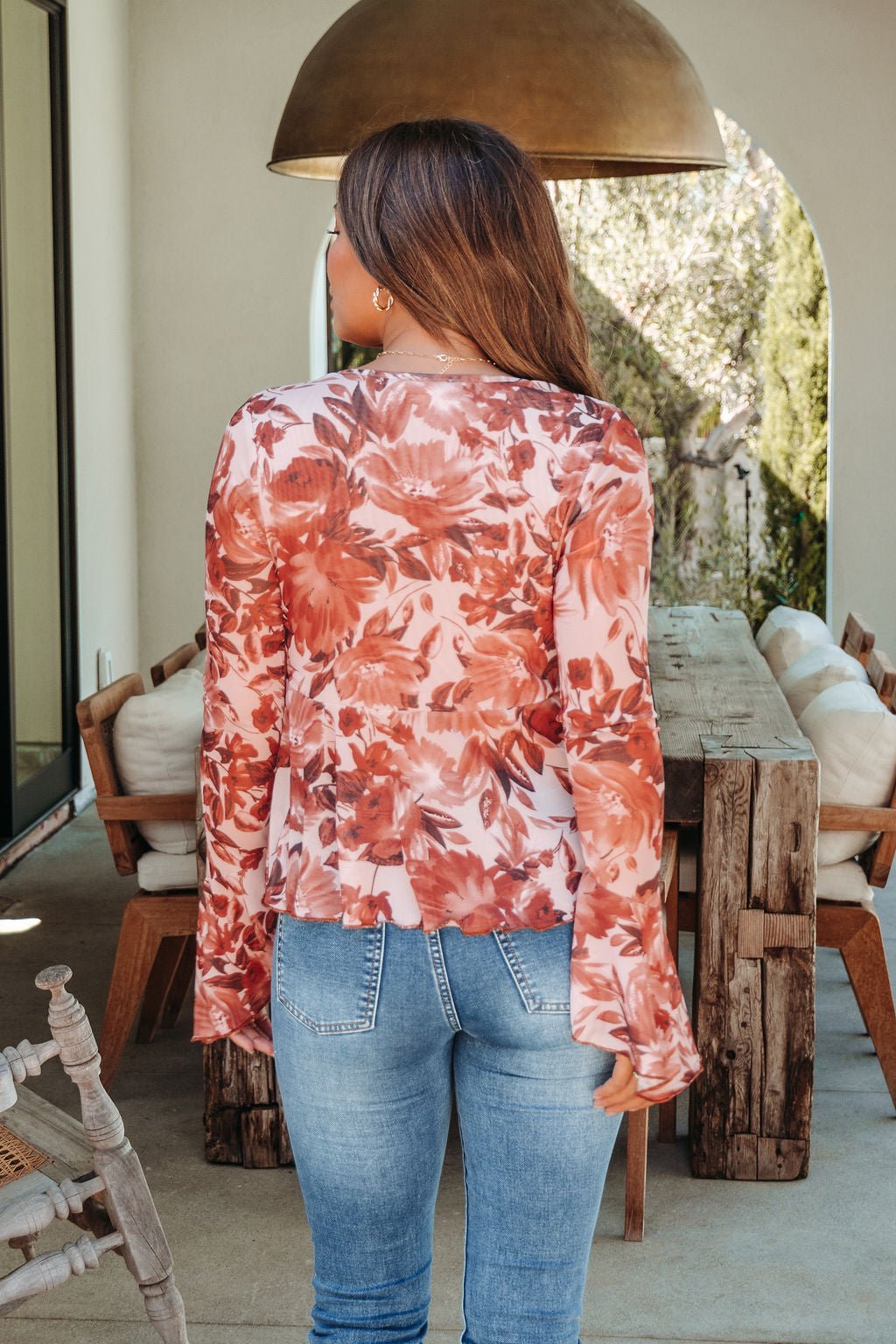 Woman with long brown hair wears the Multi Brown Floral Mesh Top and blue jeans, standing on a patio facing away from the camera.