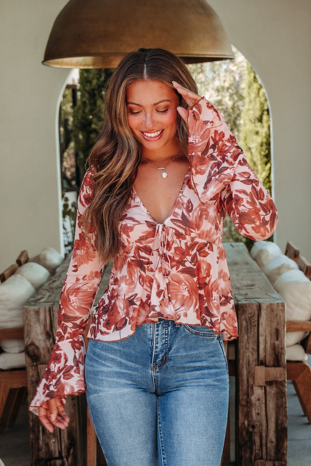 Woman in a Multi Brown Floral Mesh Top and jeans stands indoors by a rustic wooden table, touching her hair and looking down.