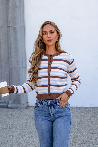 Woman in Multi Brown Striped Button Down Cardigan and jeans, holding a coffee, stands by a gray stone wall and white background.