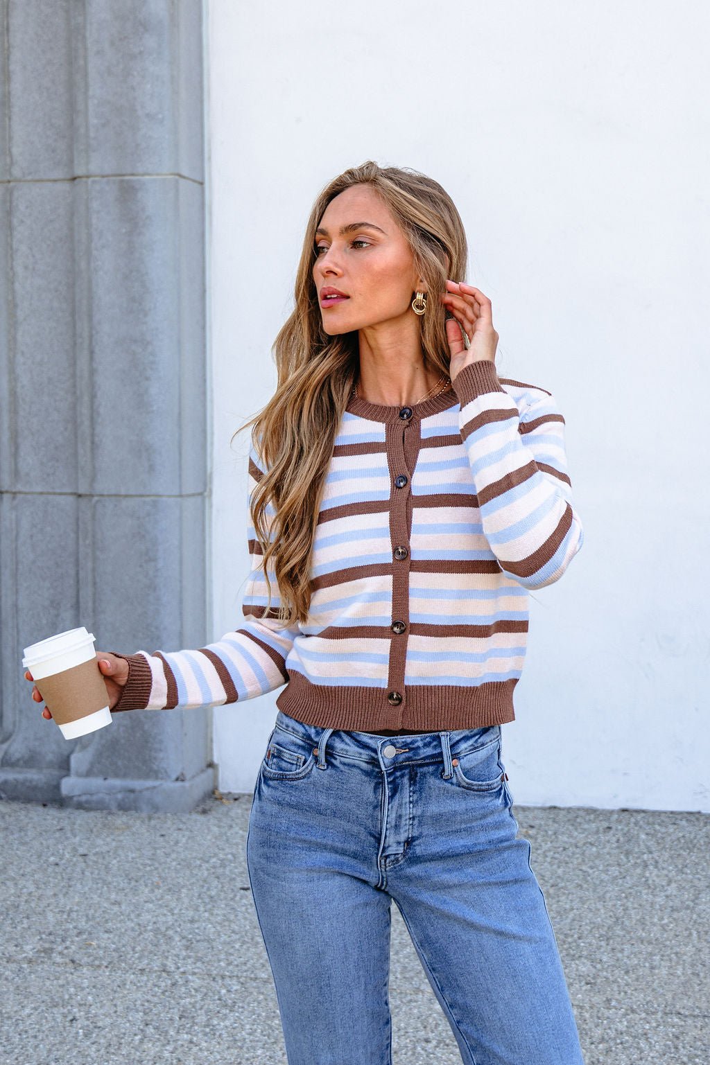 Woman in a Multi Brown Striped Button Down Cardigan and jeans, holding coffee outdoors by a gray column and white wall.