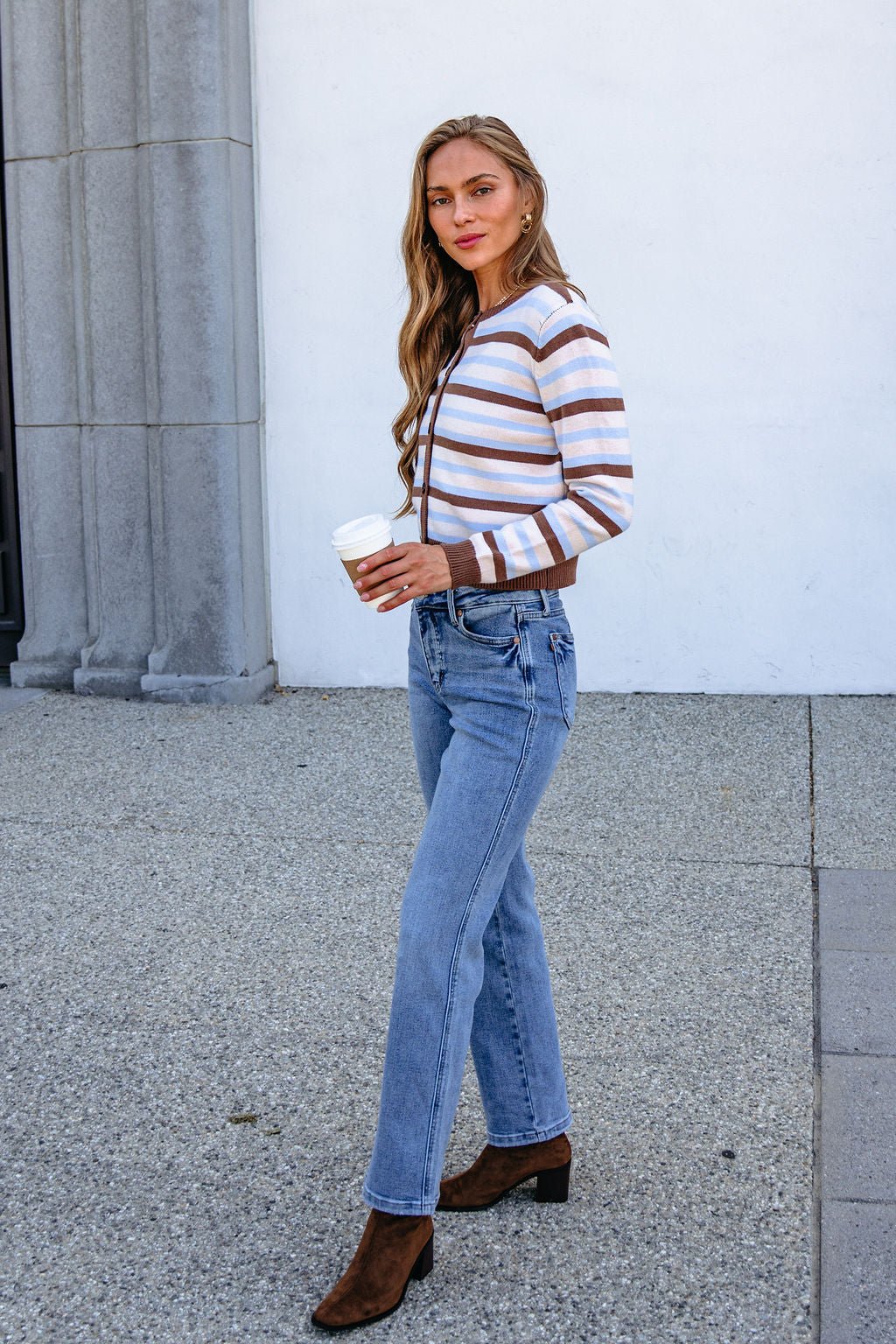 Woman in a Multi Brown Striped Button Down Cardigan, blue jeans, and boots holds coffee on a sidewalk near columned building.
