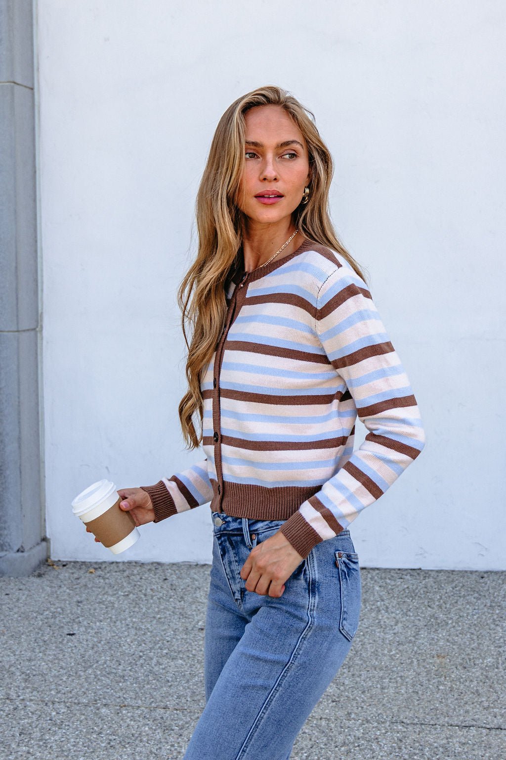 Woman in a Multi Brown Striped Button Down Cardigan and jeans, holding coffee, walks outside by a white wall.
