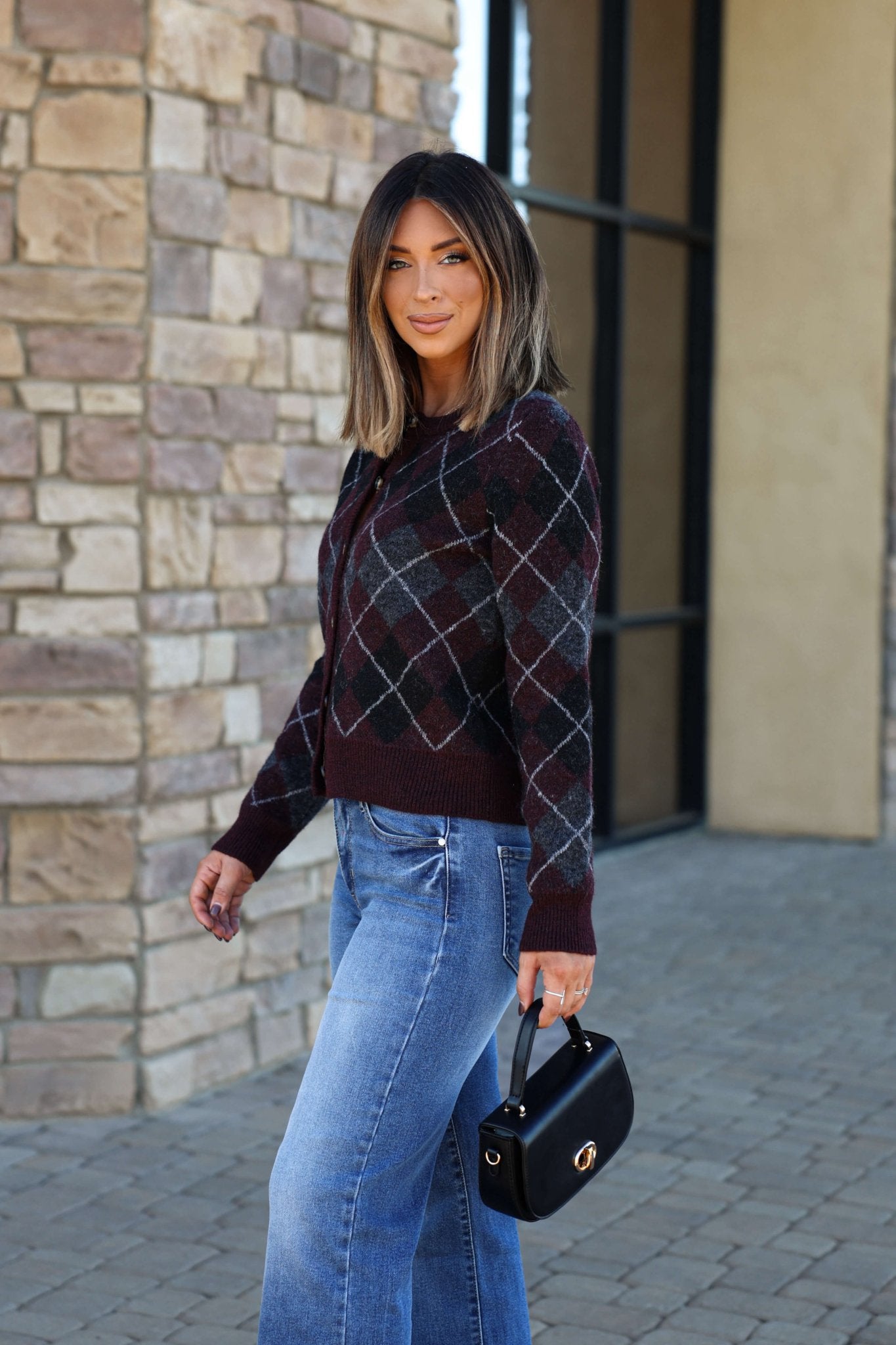 A woman wears the Multi Burgundy Argyle Knit Cardigan with jeans, holding a small black handbag and smiling outdoors.
