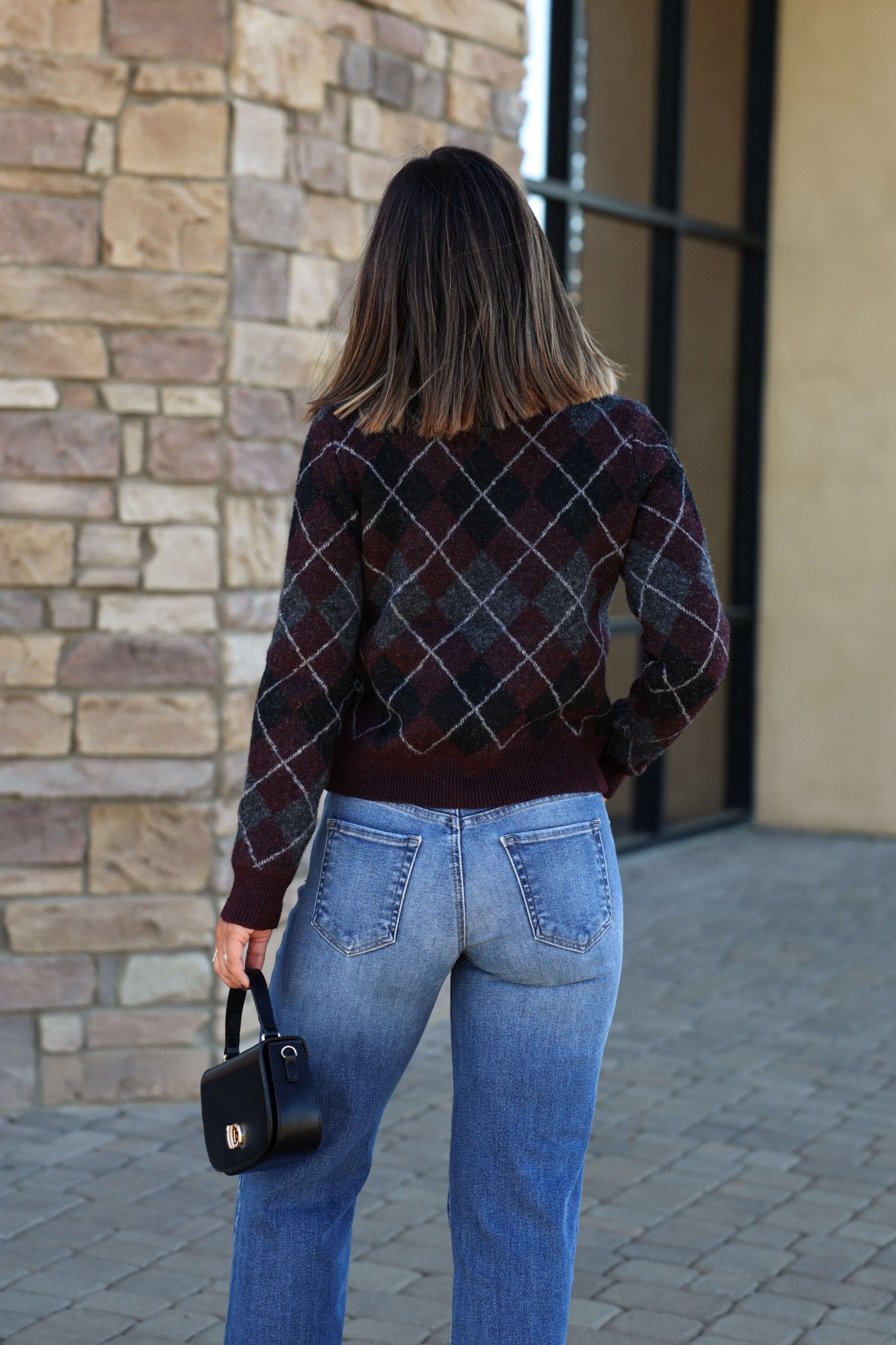 Woman in the Multi Burgundy Argyle Knit Cardigan and jeans stands outdoors, facing away, holding a small black purse.