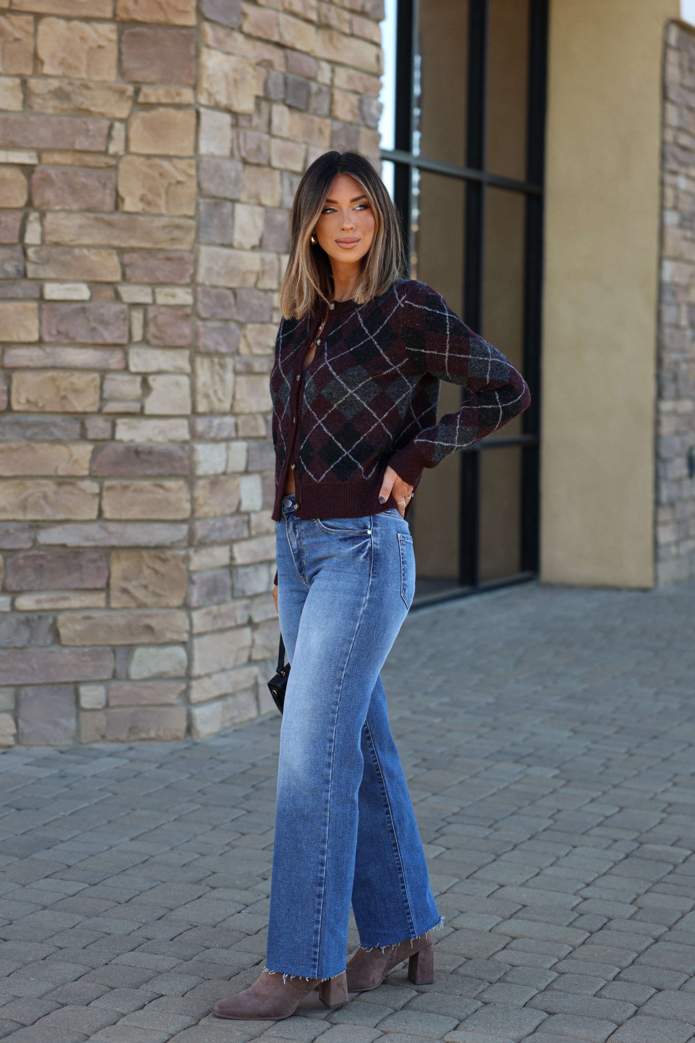 Woman in a Multi Burgundy Argyle Knit Cardigan, blue wide-leg jeans, and boots stands by a stone building on a cobblestone path.