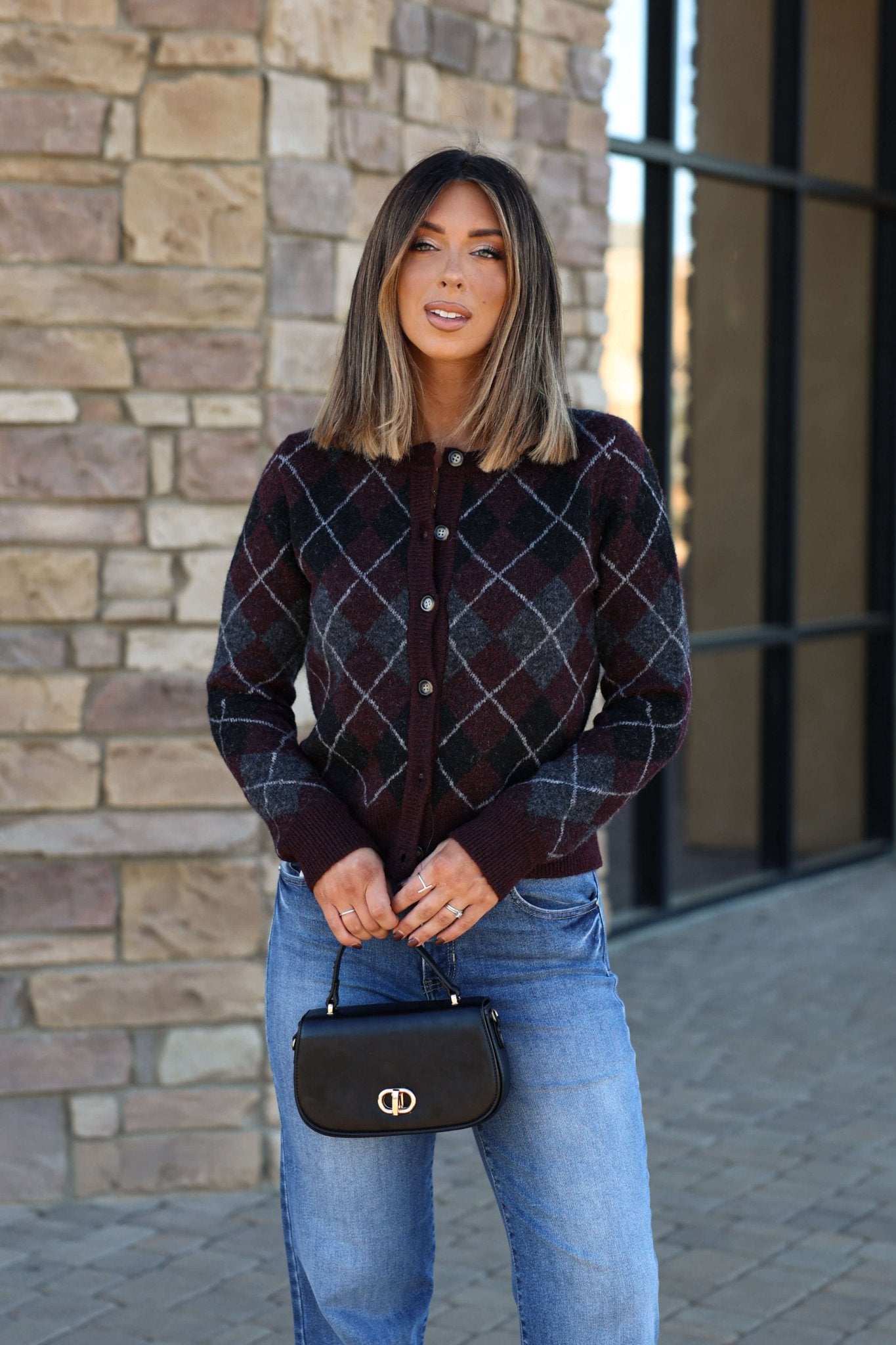 A woman with straight hair wears the Multi Burgundy Argyle Knit Cardigan, blue jeans, and holds a small black handbag outside.