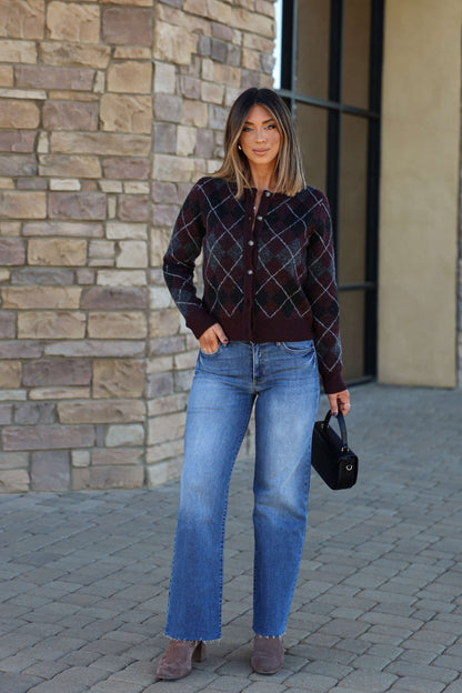 Woman with straight hair wears the Multi Burgundy Argyle Knit Cardigan, blue jeans, taupe shoes, and holds a black purse outdoors.