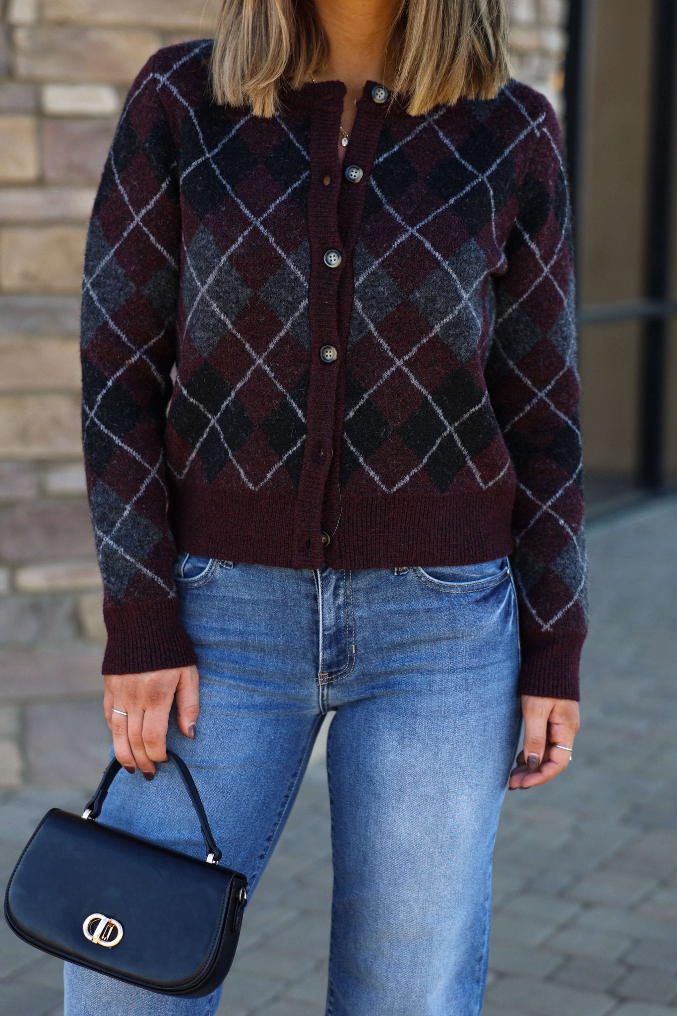 Woman wears a Multi Burgundy Argyle Knit Cardigan, blue jeans, and holds a small black handbag outside.