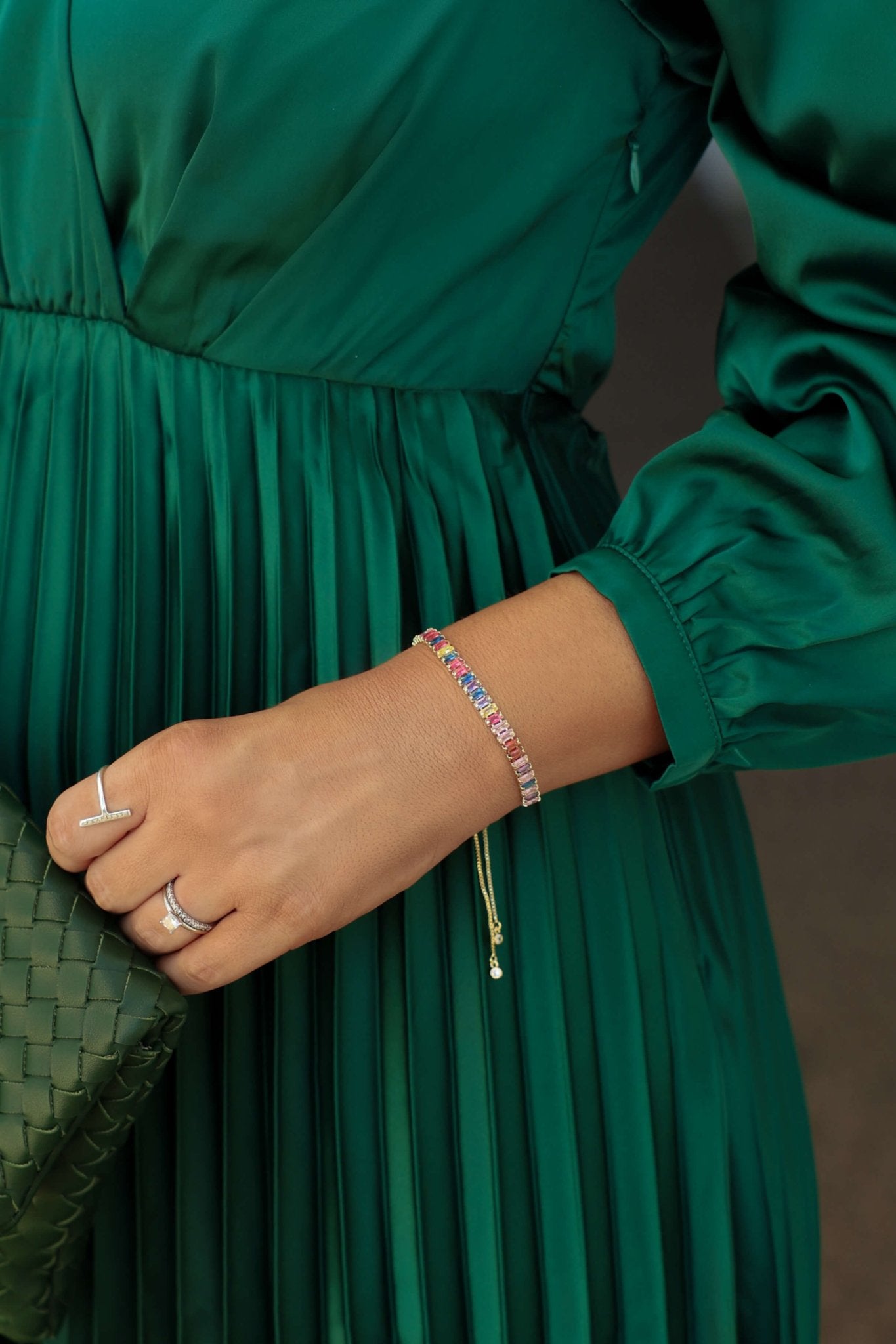 A person in a green pleated dress holds a woven clutch, wearing silver rings and the Multi Color Gem Bracelet.