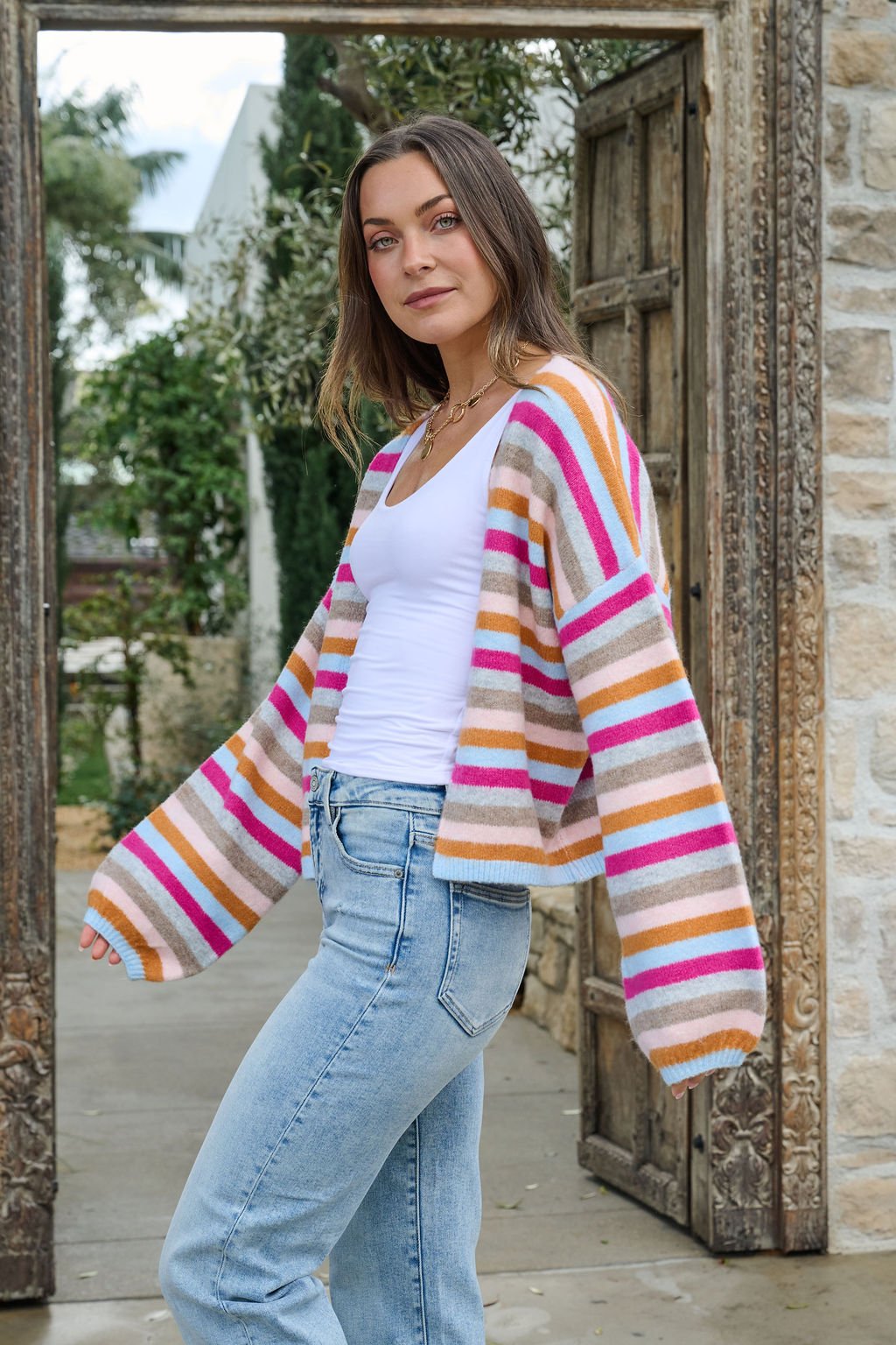 A woman in a Multi Color Striped Open Front Cardigan and jeans stands outdoors by a rustic wooden door, looking at the camera.