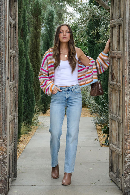 Woman wears jeans, white tank, and our Multi Color Striped Open Front Cardigan, standing in a garden doorway holding the doors open.
