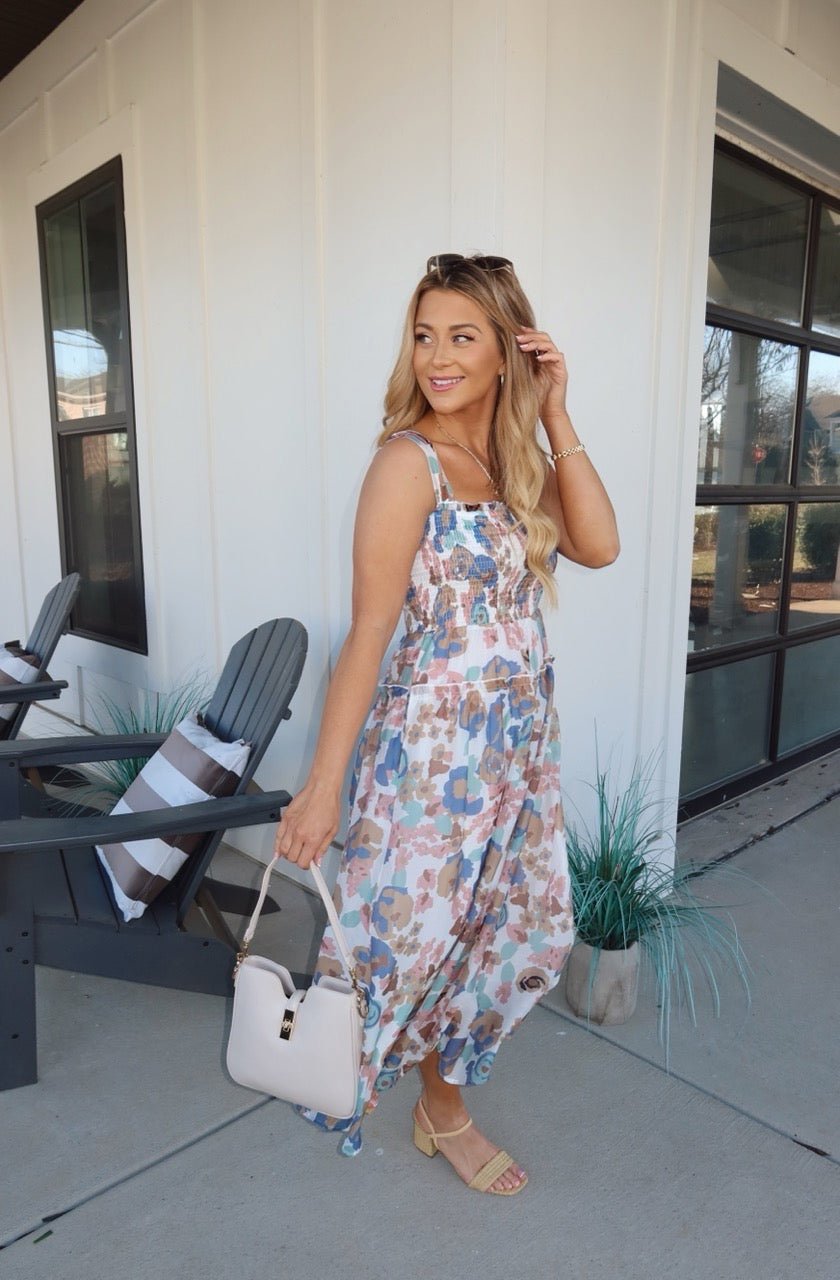 A woman wears the Multi Floral Print Smocked Midi Dress and holds a white handbag on a patio near black chairs and large windows.