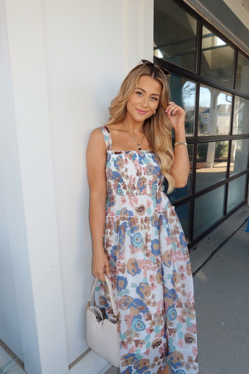 A woman smiles outside by a window, wearing the Multi Floral Print Smocked Midi Dress and holding a light-colored handbag.