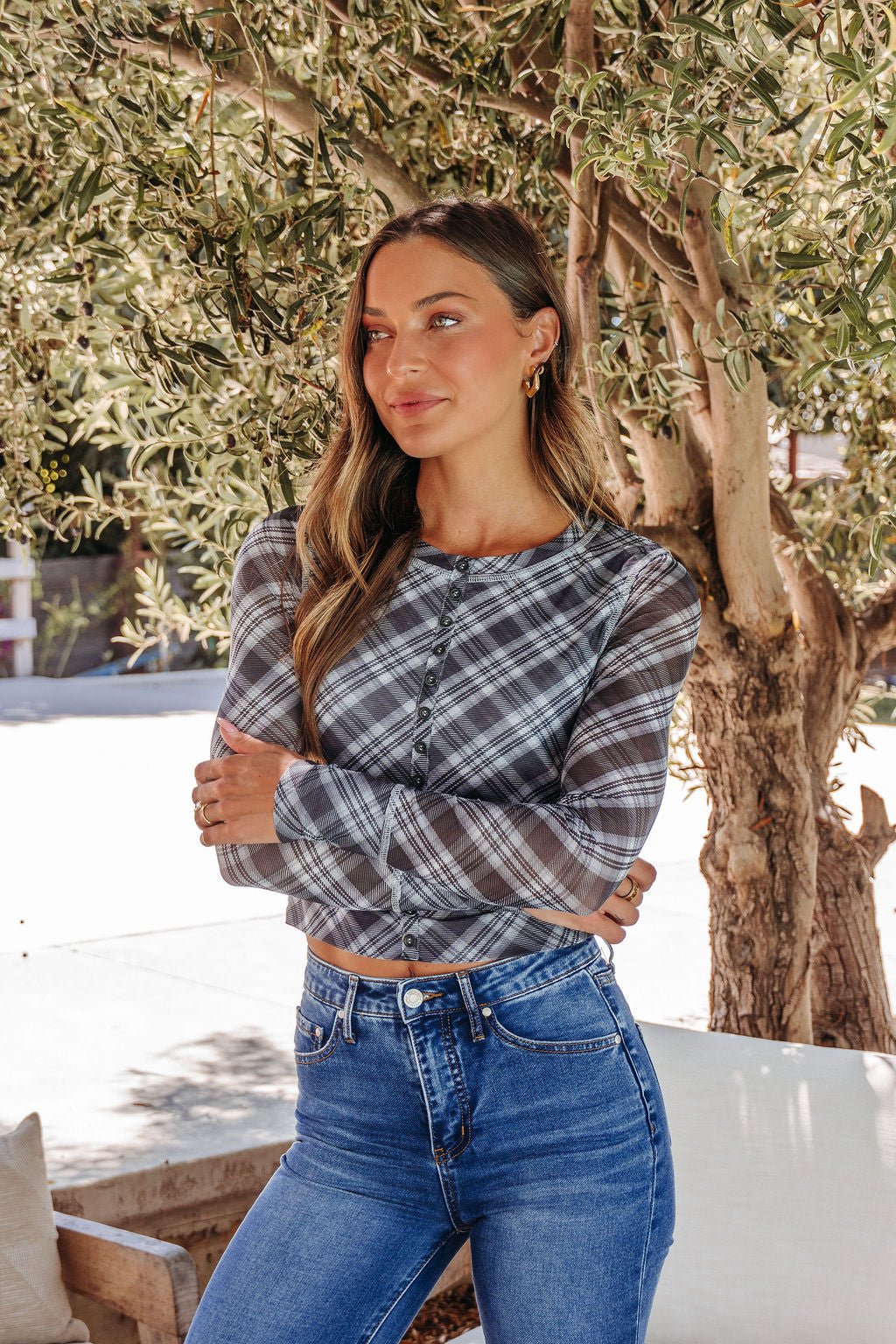 A woman with long hair wears the Multi Grey Plaid Mesh Top - DOORBUSTER and blue jeans, standing outdoors by a tree.