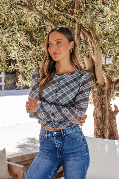 A woman with long hair wears the Multi Grey Plaid Mesh Top - DOORBUSTER and blue jeans, standing outdoors by a tree.