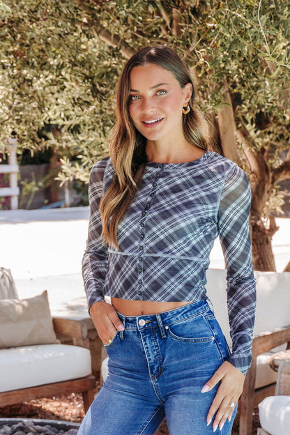 A woman wears the Multi Grey Plaid Mesh Top - DOORBUSTER with blue jeans, smiling outdoors by a tree and seating area.