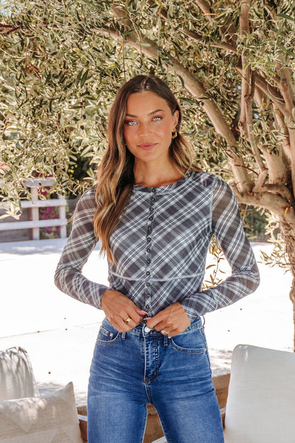 A woman wears the Multi Grey Plaid Mesh Top - DOORBUSTER and jeans outdoors by a tree, smiling slightly at the camera.