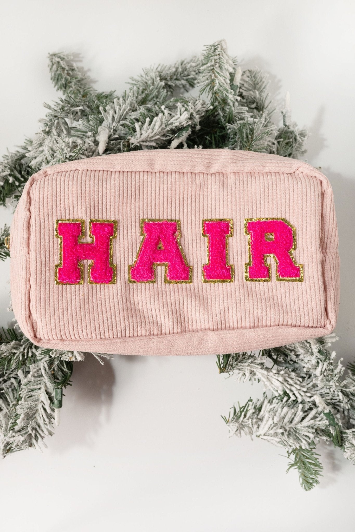 A Multi Mix Patch Make Up Bag in pink corduroy with bold pink and gold "HAIR" letters, displayed on evergreen branches.