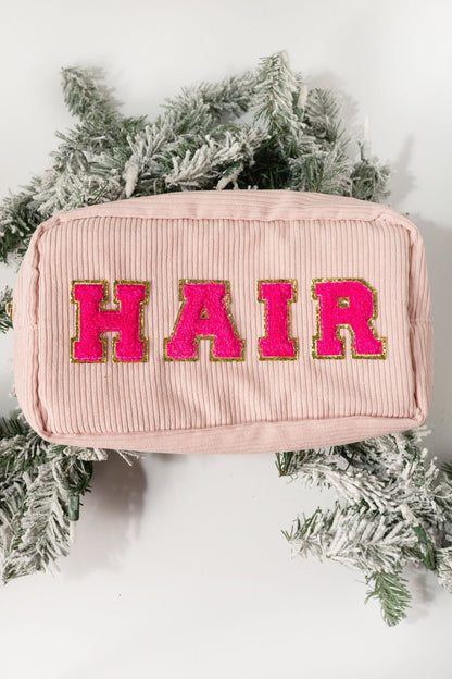 A Multi Mix Patch Make Up Bag in pink corduroy with bold pink and gold "HAIR" letters, displayed on evergreen branches.