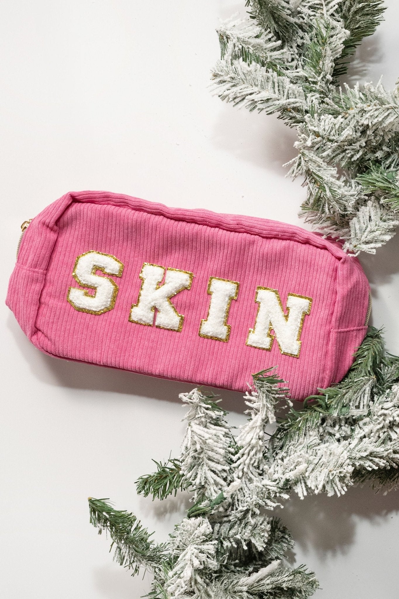 A pink Multi Mix Patch Make Up Bag with SKIN in bold white, nestled among frosted pine branches on a white background.