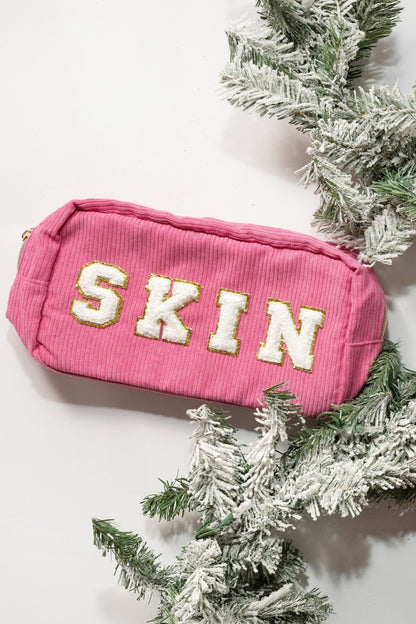 A pink Multi Mix Patch Make Up Bag with SKIN in bold white, nestled among frosted pine branches on a white background.