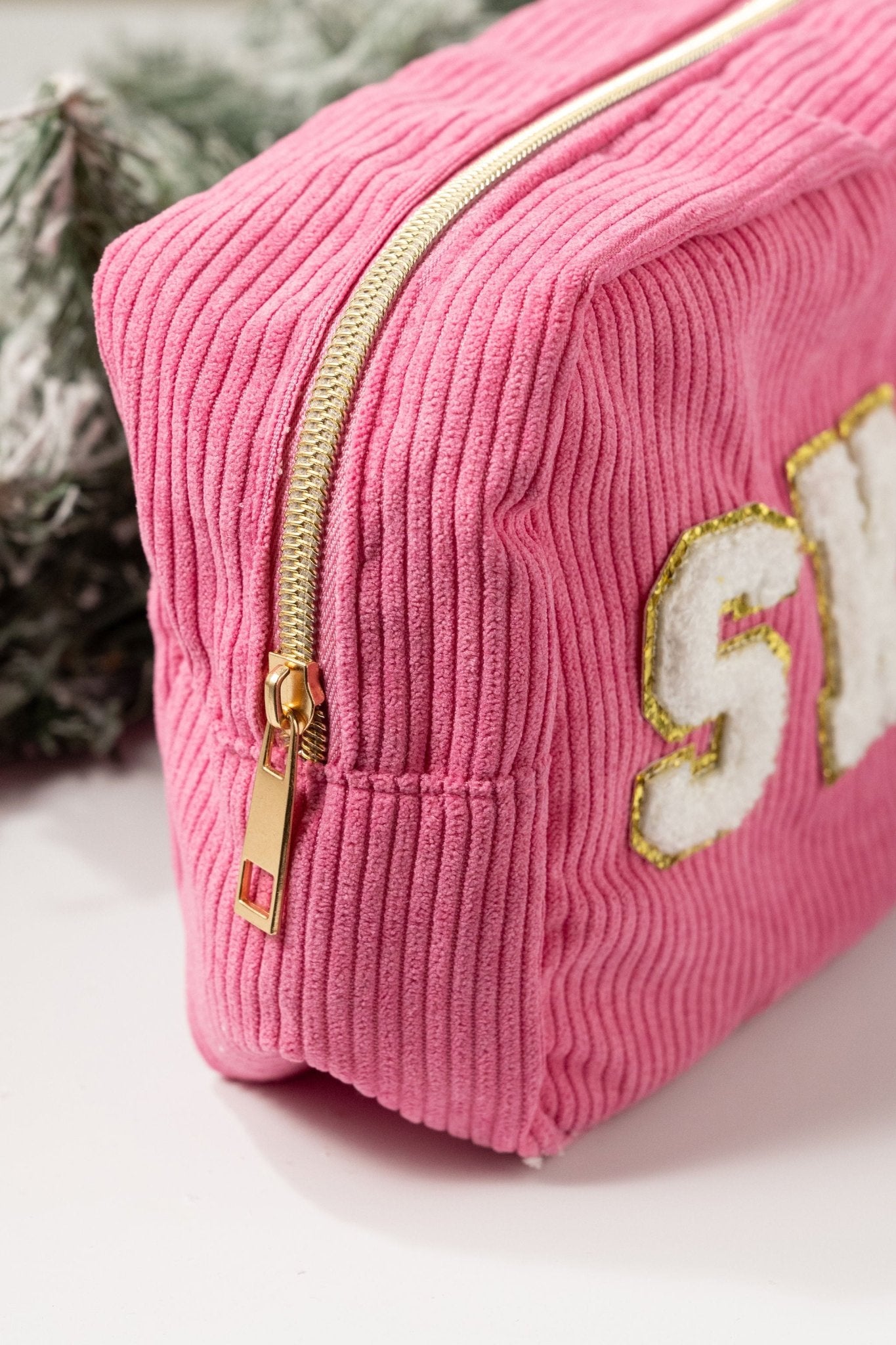 A Multi Mix Patch Make Up Bag in pink corduroy with gold trim letters, shown near green pine branches.