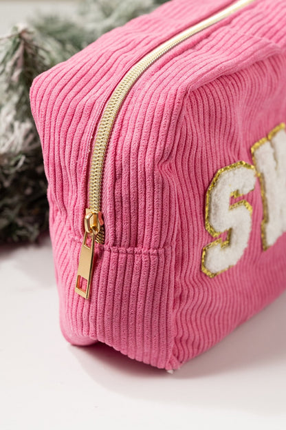 A Multi Mix Patch Make Up Bag in pink corduroy with gold trim letters, shown near green pine branches.