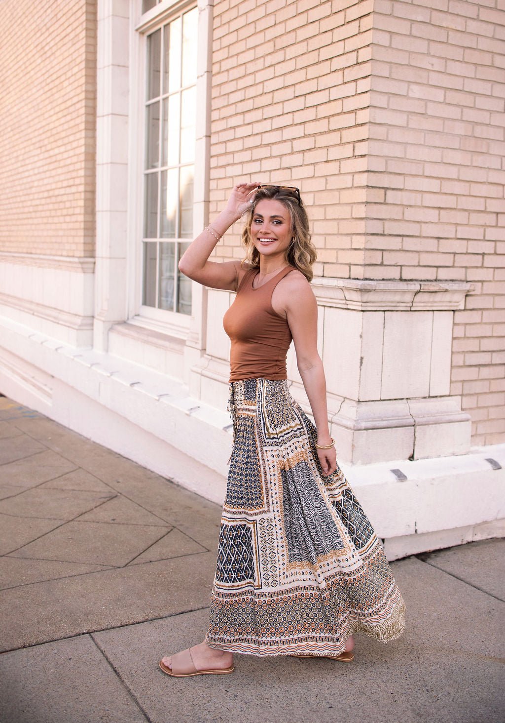 Woman smiling in a Multi Olive Print Smocked Maxi Skirt - FINAL SALE, wearing a brown sleeveless top and holding sunglasses on her head.