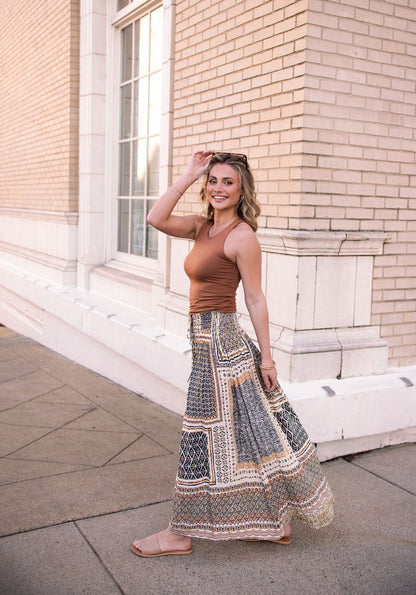 Woman smiling in a Multi Olive Print Smocked Maxi Skirt - FINAL SALE, wearing a brown sleeveless top and holding sunglasses on her head.