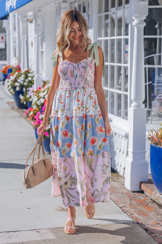 A woman in a Multi Pink Floral Bow Strap Midi Dress walks on a sunny sidewalk, holding a tan bag amid flowers and white buildings.