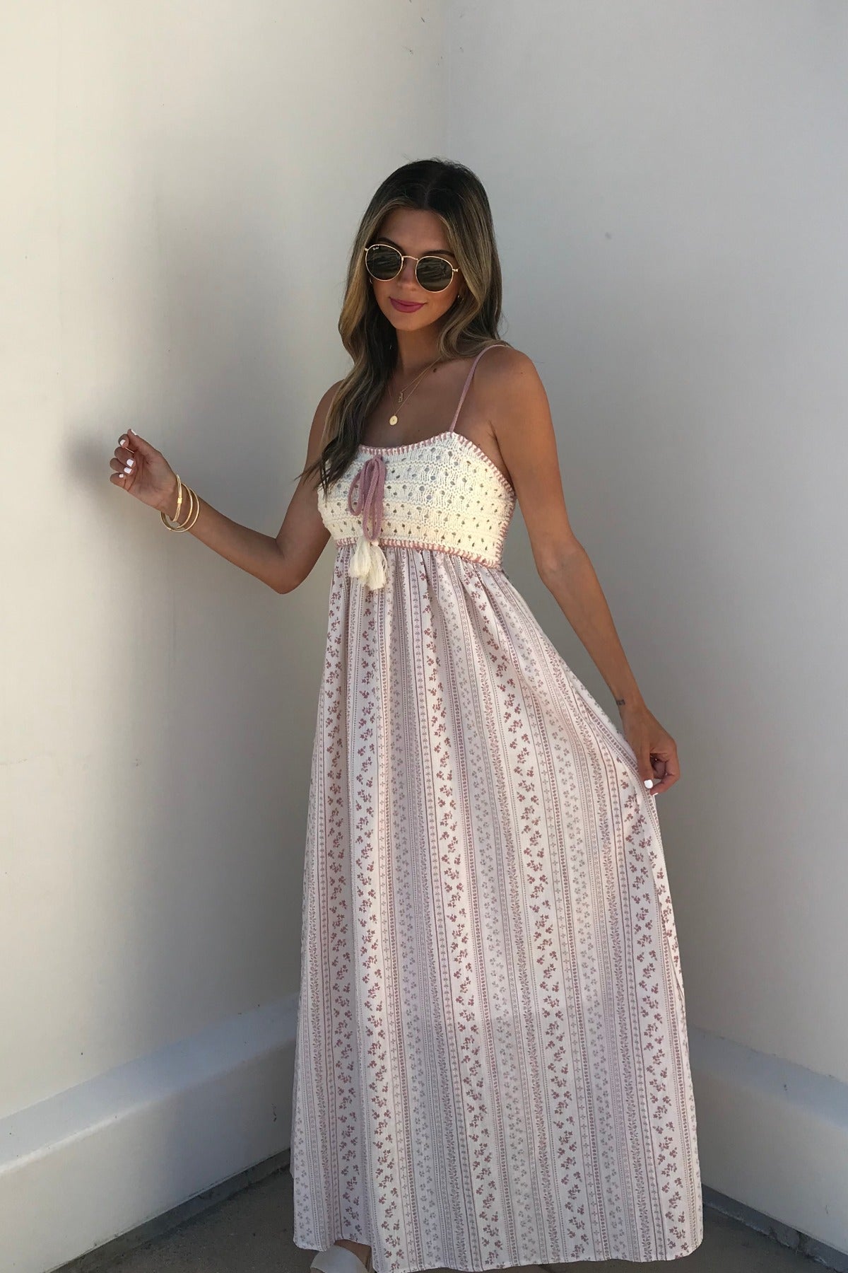 Woman wears sunglasses and a Multi Pink Stripe Crochet Maxi Dress, standing against a white wall and holding her dress out slightly.