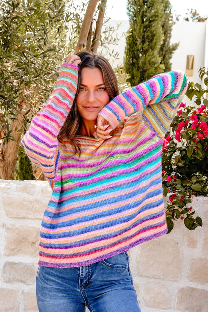 Woman smiles outdoors in a Multi Rainbow Striped Sweater and blue jeans by a stone wall surrounded by greenery.