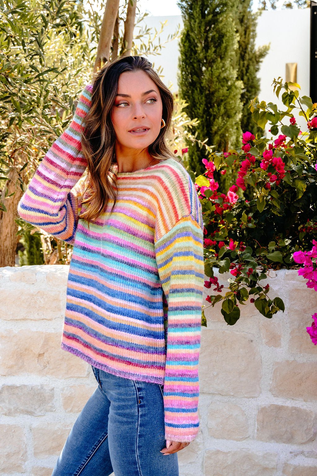 Woman in blue jeans and a Multi Rainbow Striped Sweater stands outdoors by a stone wall and flowers, looking to the side.
