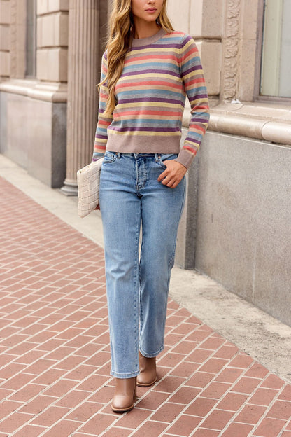 Woman in a Multi Stripe Ribbed Sweater, blue jeans, and taupe boots walks on a brick sidewalk holding a white woven clutch.