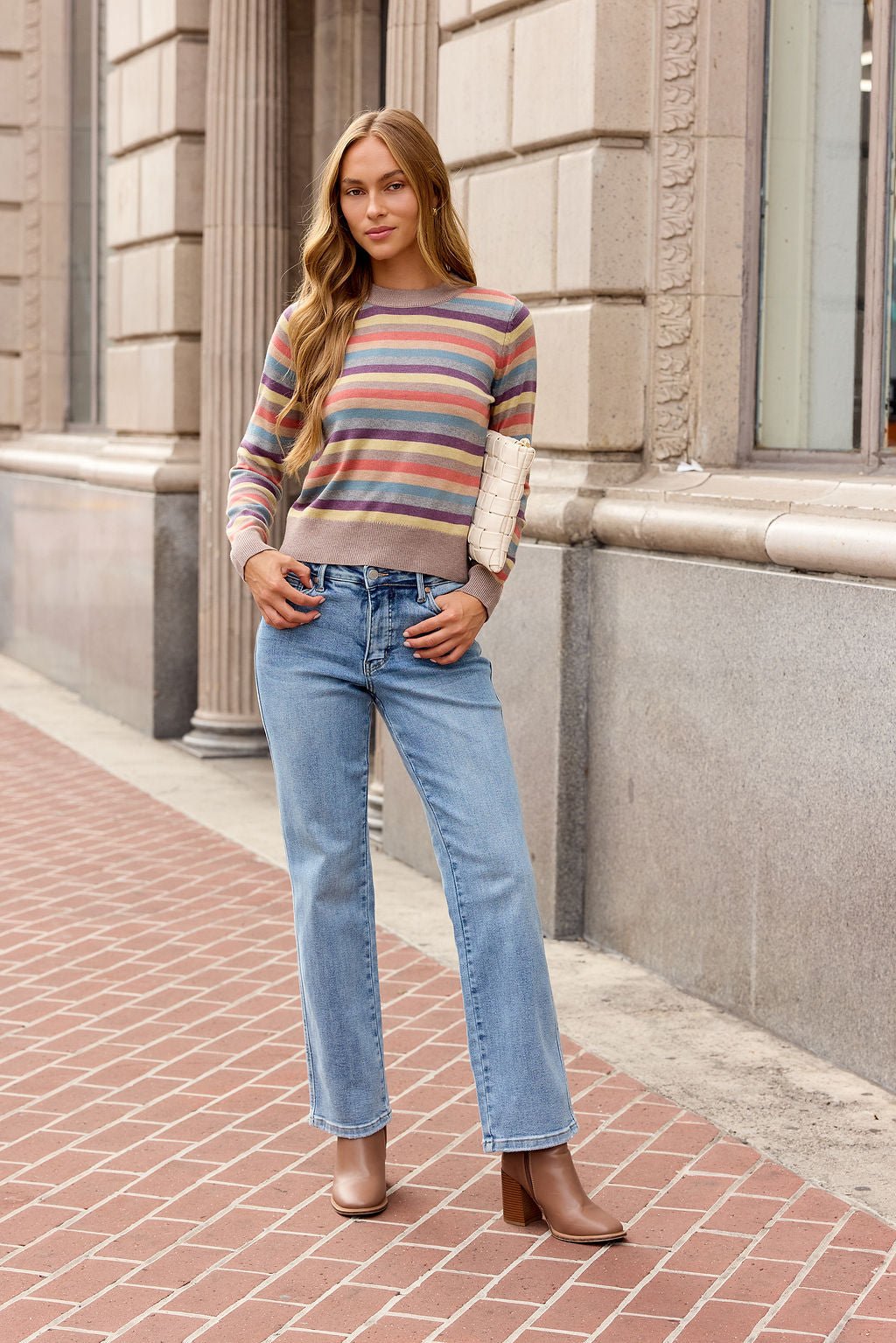 Woman wearing a Multi Stripe Ribbed Sweater, blue jeans, and brown boots stands on a brick sidewalk holding a small clutch.