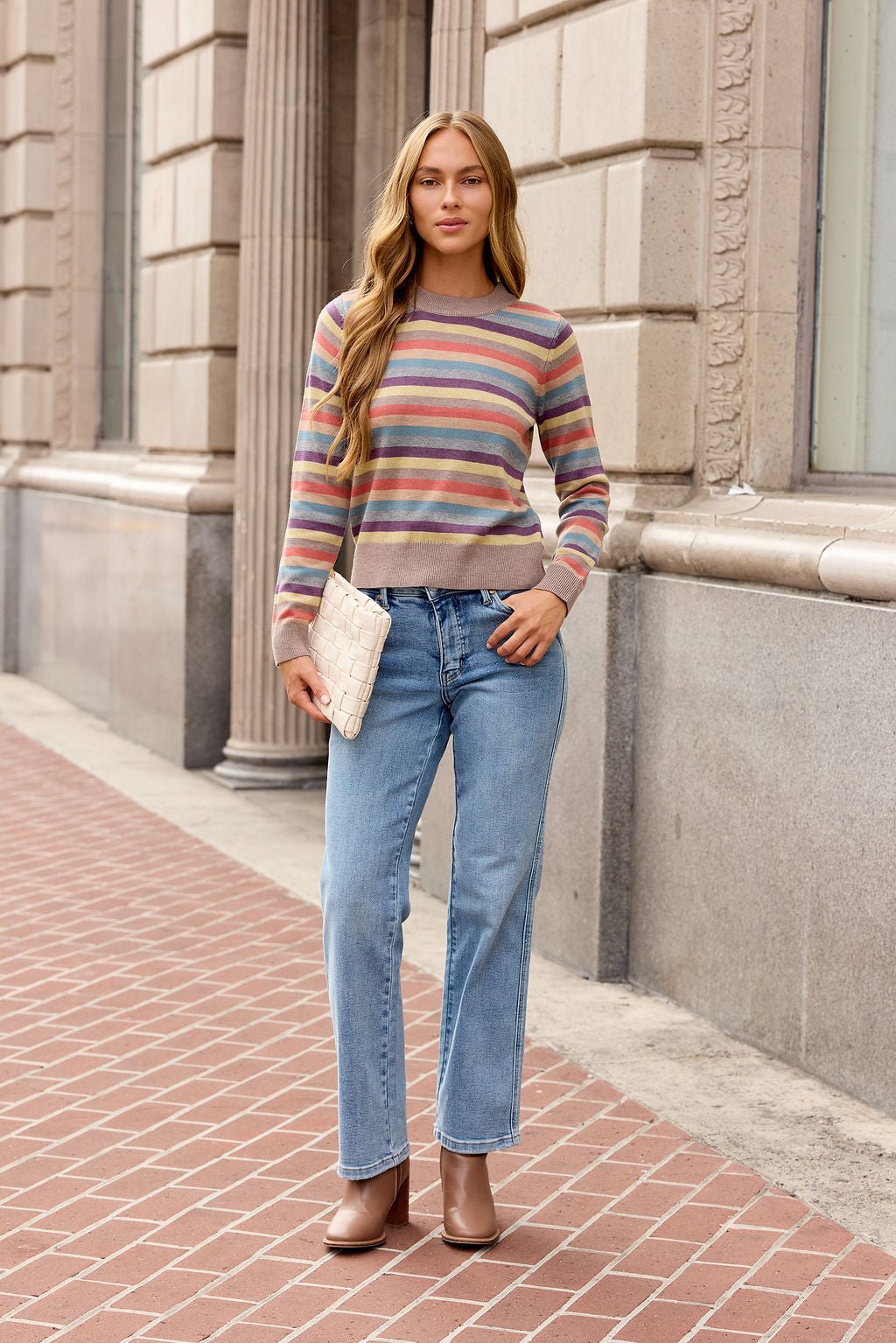 Woman in jeans and boots holds a clutch, wearing the Multi Stripe Ribbed Sweater on a brick sidewalk by a stone building.