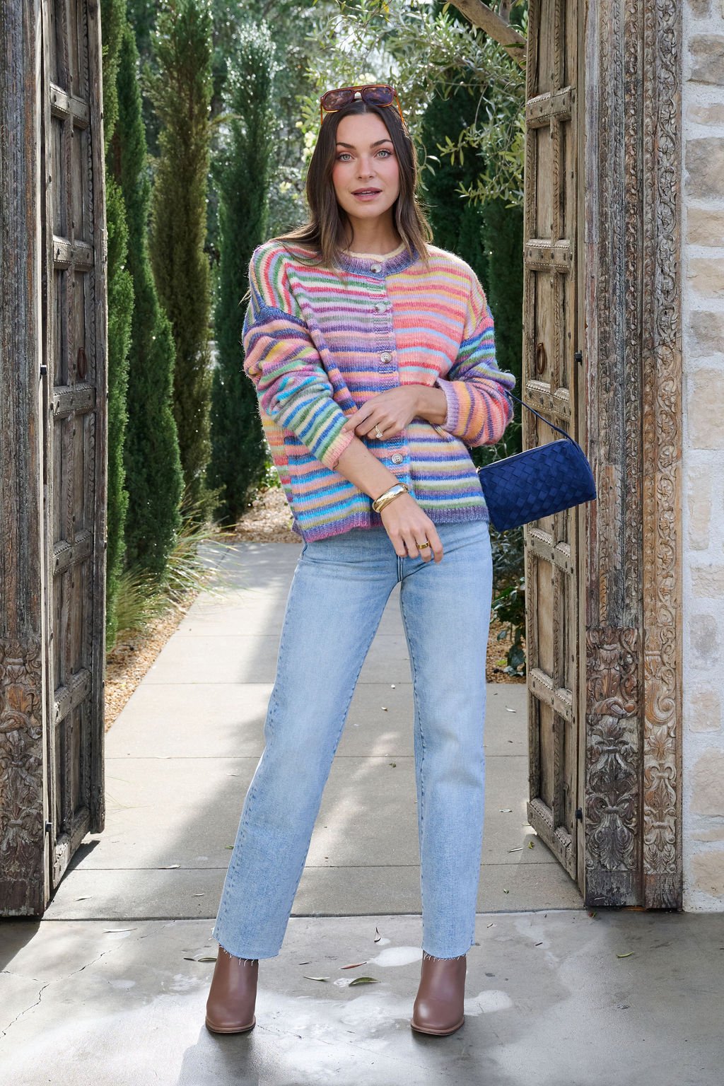 Woman outdoors between ornate doors, wearing a Multi Striped Button Down Sweater Cardigan, light blue jeans, and brown ankle boots.