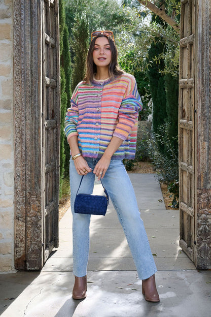 Woman in a Multi Striped Button Down Sweater Cardigan, light blue jeans, and brown boots stands outdoors with a small blue purse.