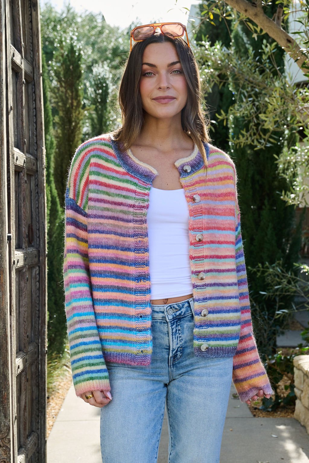 A woman in a Multi Striped Button Down Sweater Cardigan, white tank top, and jeans smiles outdoors with greenery in the background.