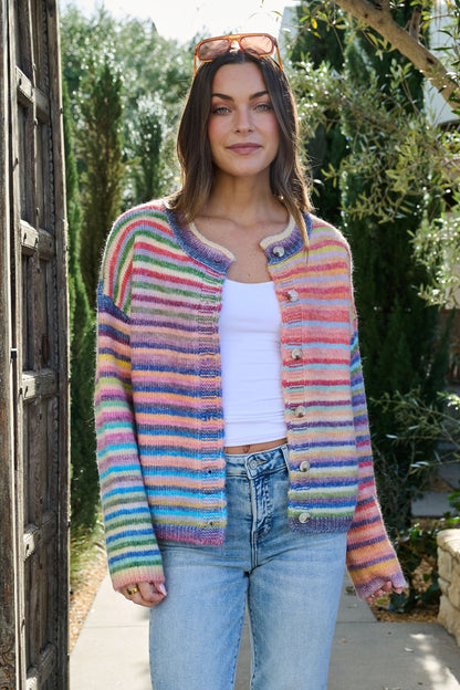 A woman in a Multi Striped Button Down Sweater Cardigan, white tank top, and jeans smiles outdoors with greenery in the background.
