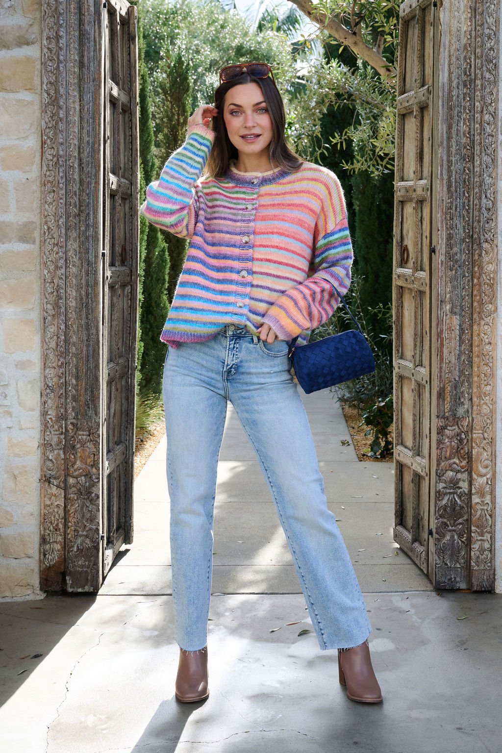 Woman in a Multi Striped Button Down Sweater Cardigan, blue jeans, and brown boots stands in a rustic doorway holding a blue bag.