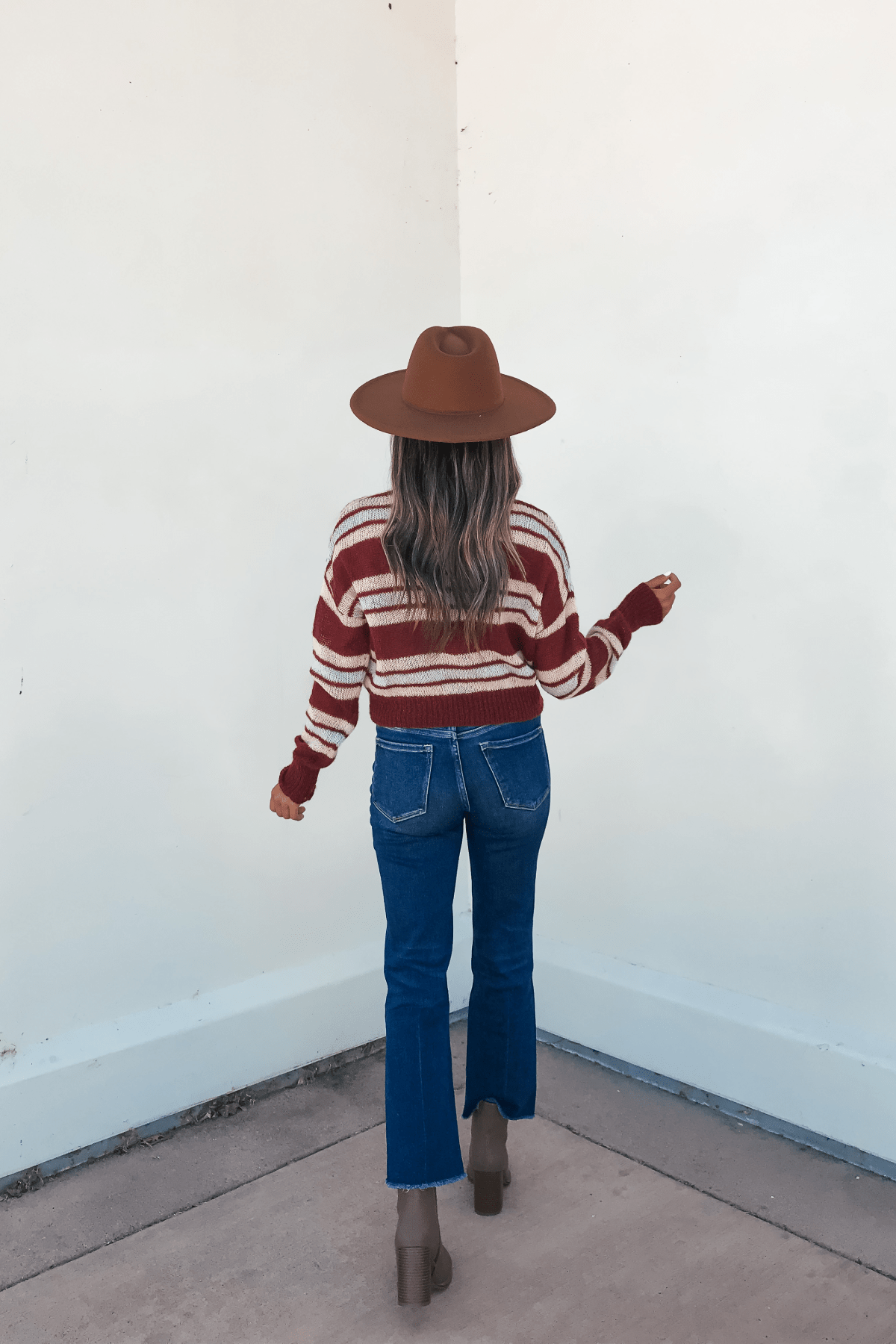 A woman in a Multi Striped Button Up Sweater Cardigan, blue jeans, and brown hat stands with her back to a white wall.
