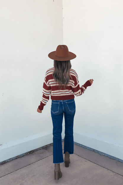 A woman in a Multi Striped Button Up Sweater Cardigan, blue jeans, and brown hat stands with her back to a white wall.