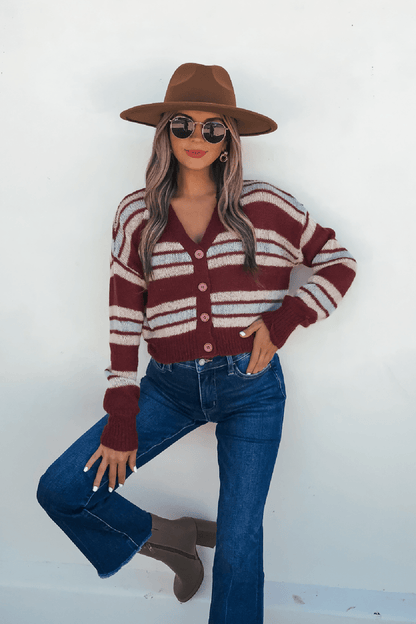 Woman in a cozy-chic Multi Striped Button Up Sweater Cardigan, wide-brim hat, jeans, and taupe boots poses by a white wall.
