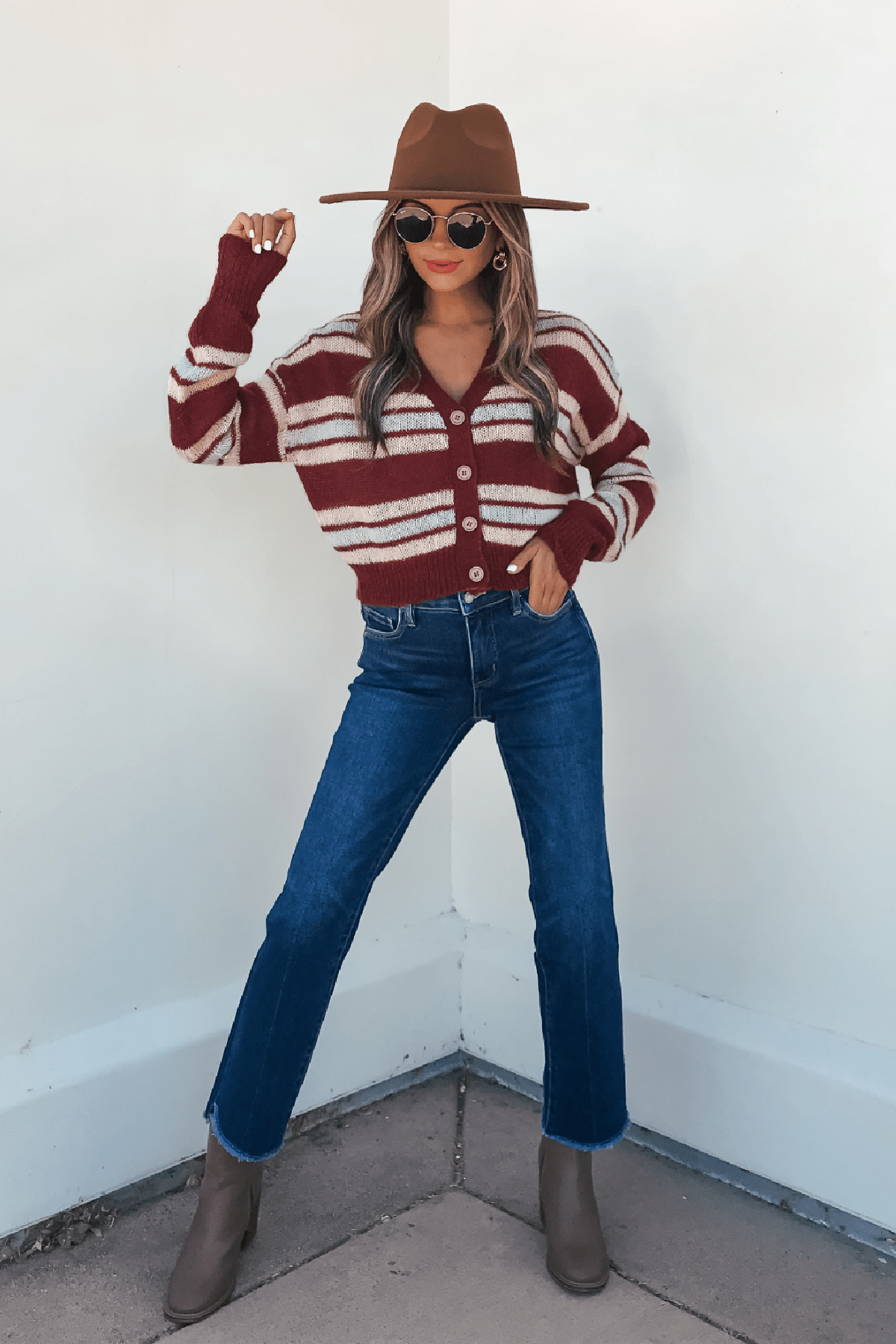 Woman in a Multi Striped Button Up Sweater Cardigan, jeans, brown hat, and boots poses confidently by a white wall.