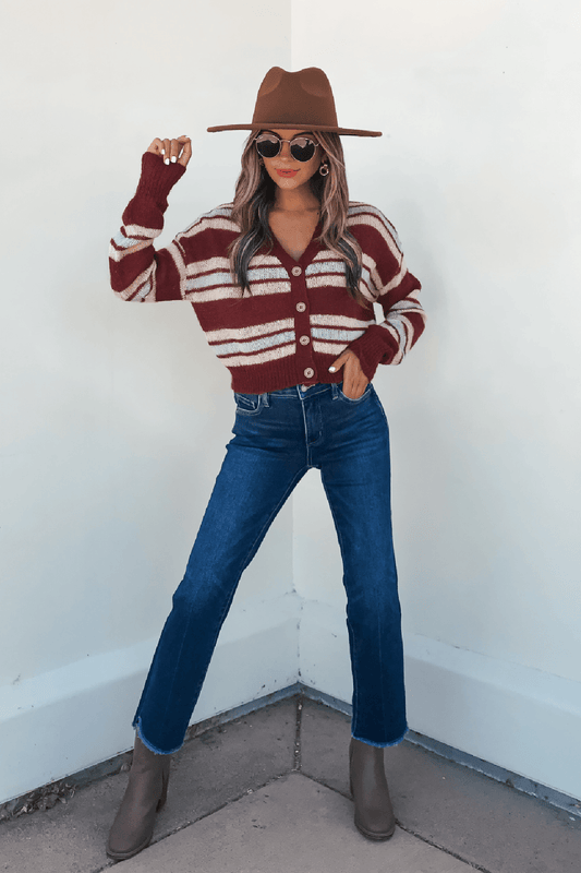 Woman wearing sunglasses, blue jeans, brown boots, and a Multi Striped Button Up Sweater Cardigan standing by a white wall.