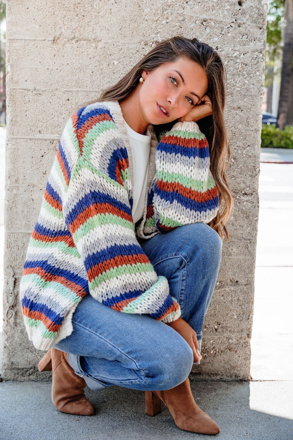 Woman in a Multi Striped Open Front Sweater Cardigan squats by a concrete wall, head resting on her hand.