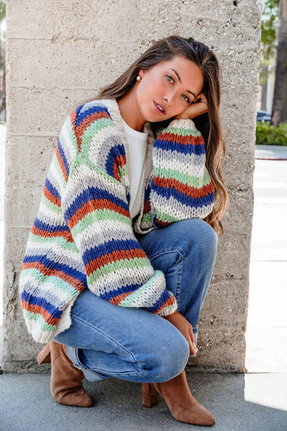 Woman in a Multi Striped Open Front Sweater Cardigan squats by a concrete wall, head resting on her hand.
