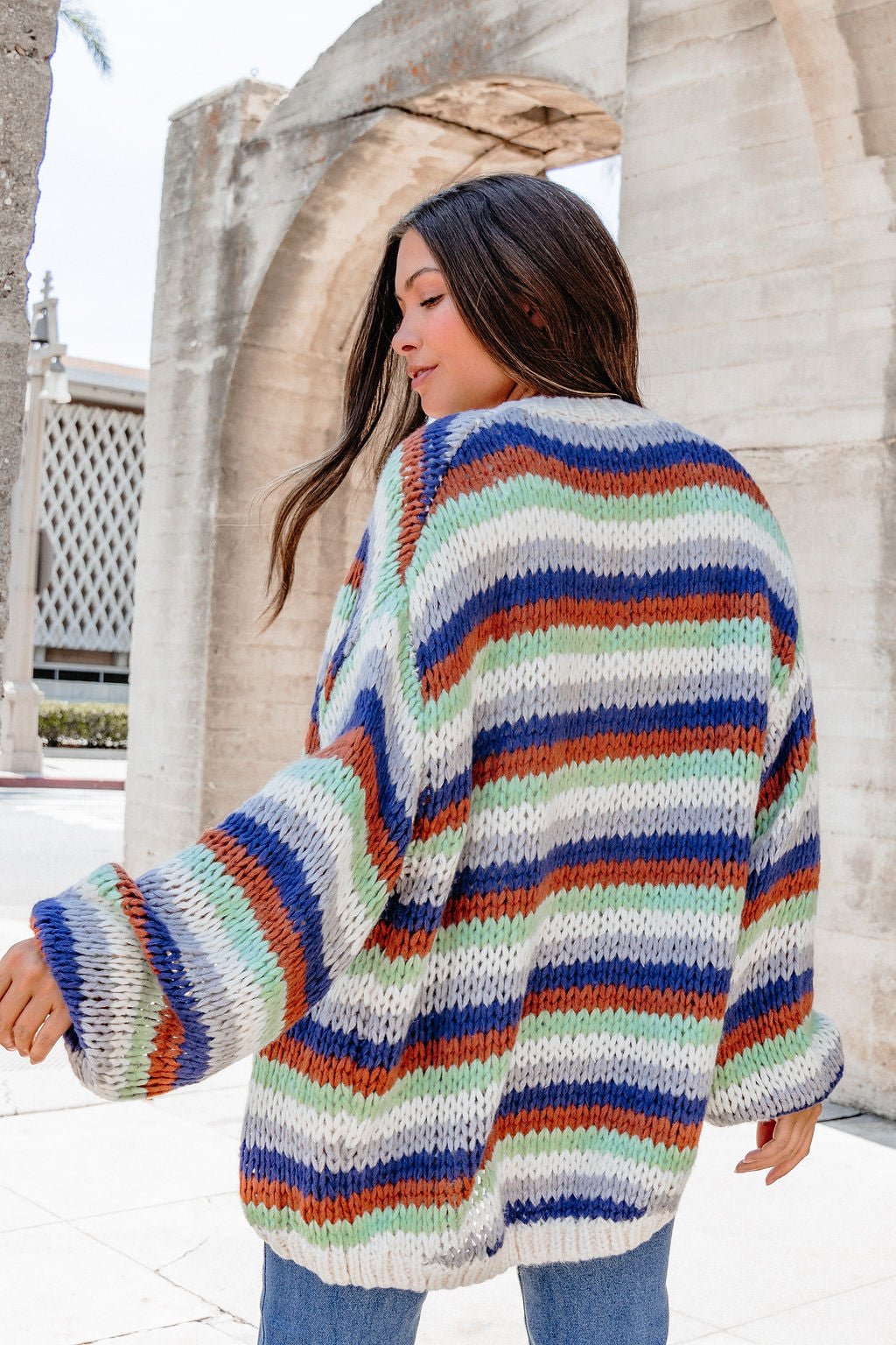 A woman models the Multi Striped Open Front Sweater Cardigan—FINAL SALE—outdoors by stone arches, facing away from the camera.